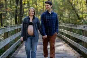 A couple takes maternity photos on a scenic wooden boardwalk surrounded by trees.