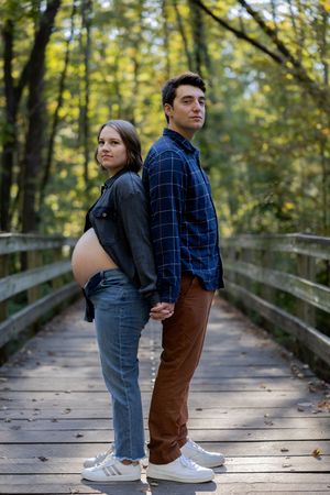 A couple poses back-to-back on a wooden walkway in an autumn forest setting.