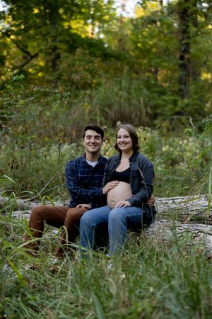 A couple shares joyful moments during their maternity photoshoot in a natural setting.