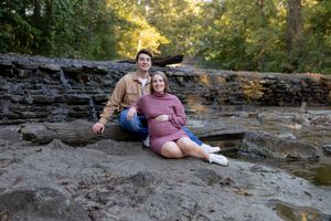 A couple relaxes on rocky terrain near a creek surrounded by trees.
