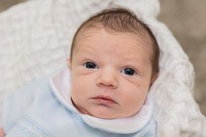 Newborn baby wearing white outfit rests on textured white blanket.