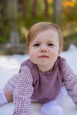 Baby in purple gingham outfit sitting on white blanket in autumn setting.