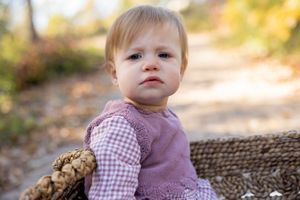 Baby wearing purple dress and gingham shirt sitting by woven basket outdoors.