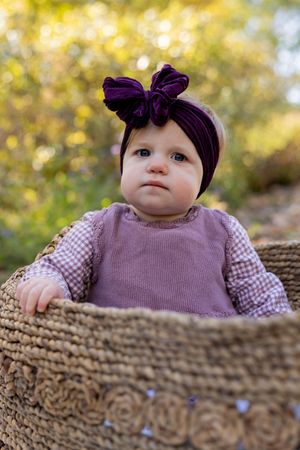 Baby wearing purple sweater and bow headband peeks over textured surface in autumn setting.