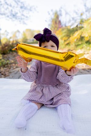 Baby in checkered dress plays with yellow blanket on white surface.
