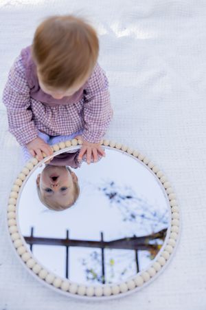 Baby explores round mirror with wooden frame placed on white surface.