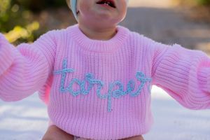 Baby wearing pink knit sweater with text detail smiles outdoors.