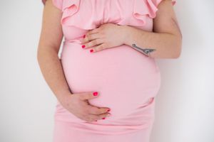 A pregnant woman in a pink dress holds her belly with red painted nails against a white background.