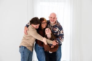 A family of four hugs and laughs together while posing against a white wall in casual clothing.