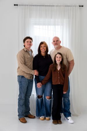 A family of four poses together in casual clothing against a white backdrop with sheer curtains.