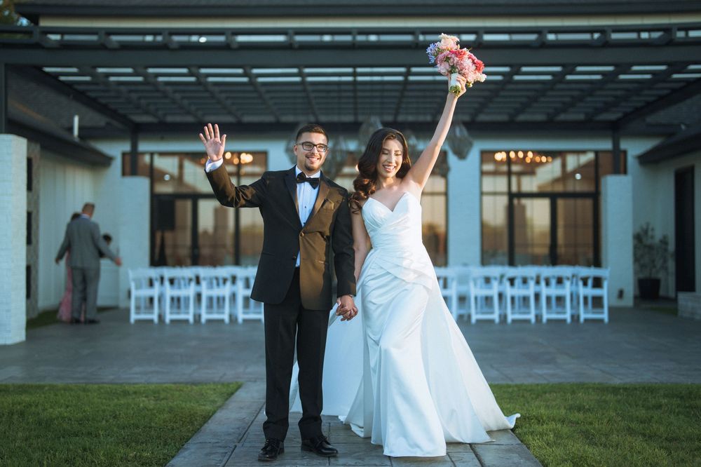 A joyful wedding couple celebrates with raised arms in front of rows of white chairs at an outdoor venue.