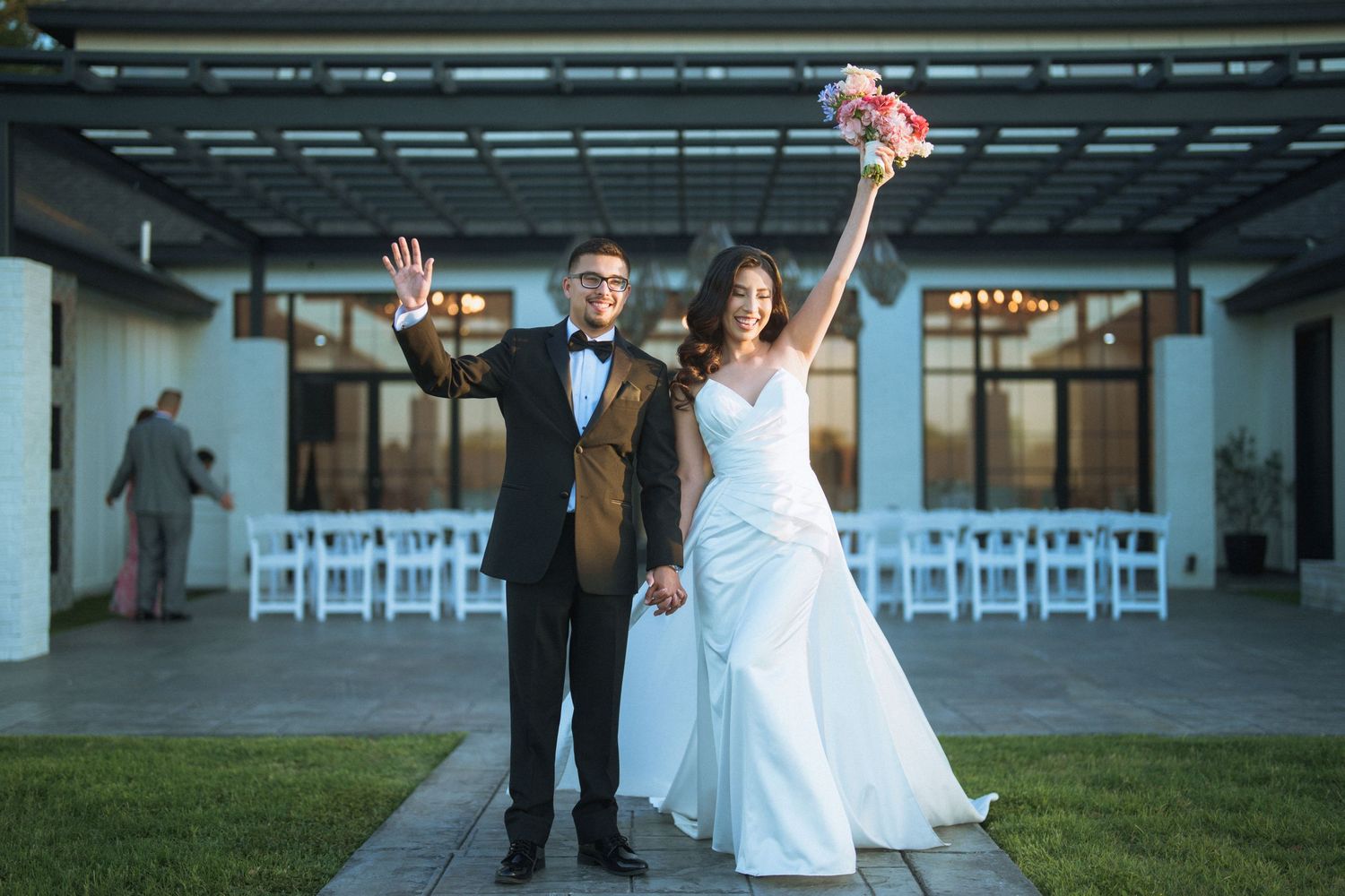 A joyful wedding couple celebrates with raised arms in front of rows of white chairs at an outdoor venue.