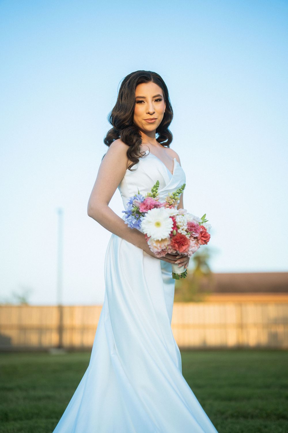 A bride in a fitted white gown holds a vibrant bouquet against a blue sky.