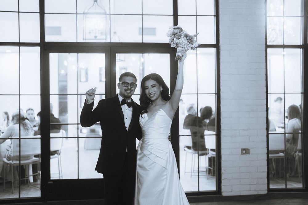 Black and white photo of newlyweds celebrating with raised arms in front of large windows.