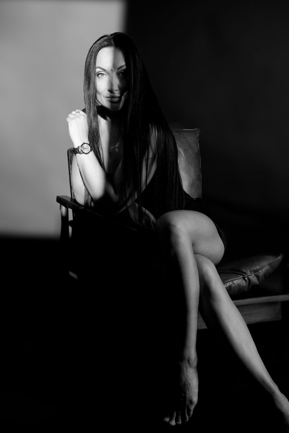 Black and white portrait of a woman with long dark hair sitting on a chair, wearing a short dress and a wrist watch.