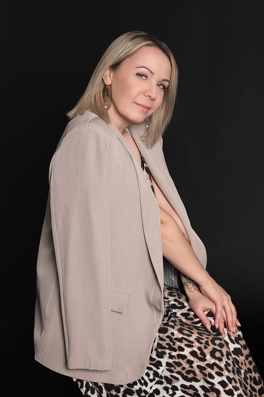 Blonde woman in beige blazer and leopard print skirt poses confidently against a dark studio background.