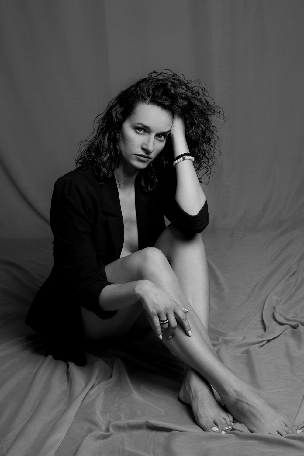 Black and white portrait of a woman with curly hair sitting on fabric, wearing a blazer and jewelry.