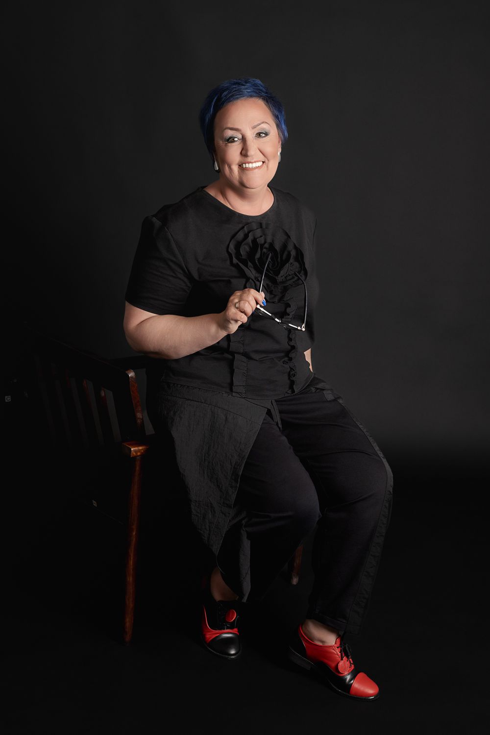 Smiling person with blue hair wearing all black outfit and red shoes, seated on chair against dark background.