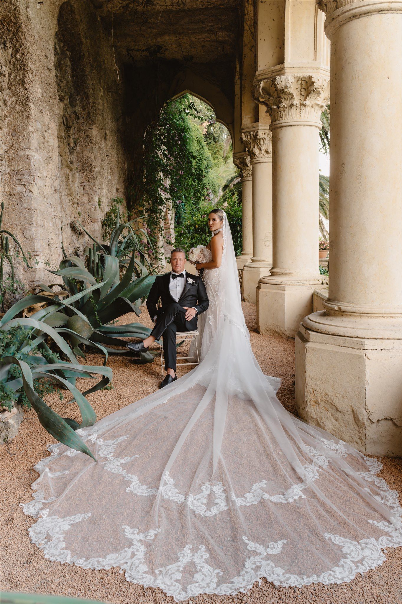 A wedding dress with intricate lace train displayed in a historic Italian villa colonnade.