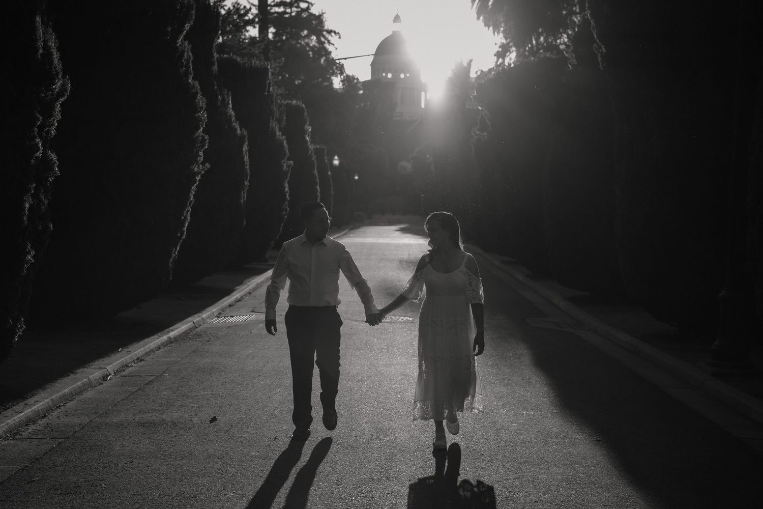 Silhouettes of couple holding hands while walking down a tree-lined path at sunset in black and white.