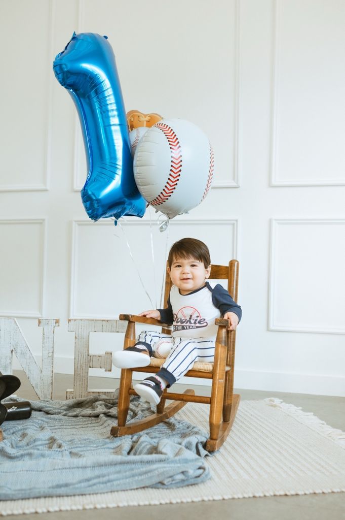 Baseball themed photo shoot setup with a wooden rocking chair, blue number 1 balloon and baseball balloon on white backdrop.