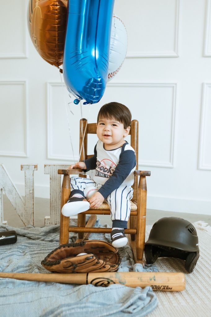 Baseball equipment and wooden rocking chair arranged with blue balloon decorations for first birthday shoot.