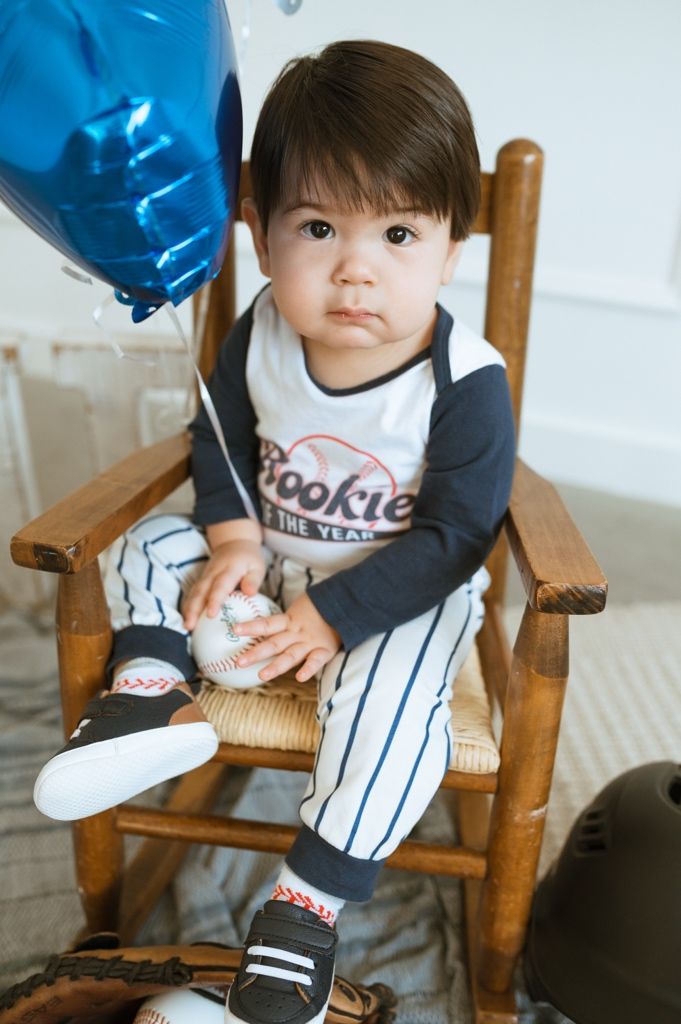 Small child seated in wooden chair with baseball themed decorations and blue balloons for a special occasion.