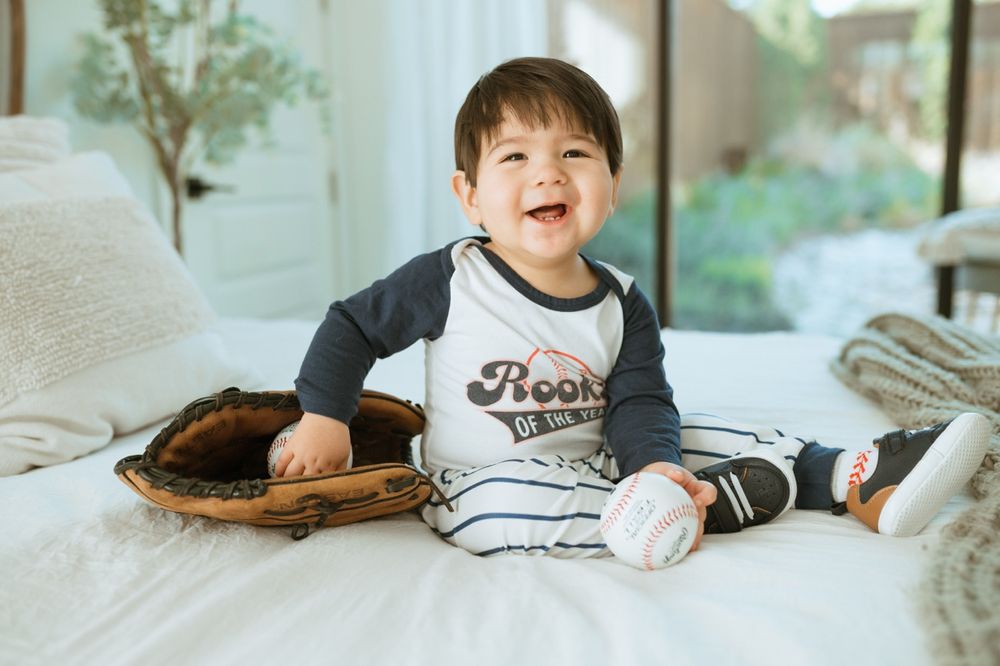 Happy toddler in baseball-themed outfit sits on white bed with baseball glove and ball.