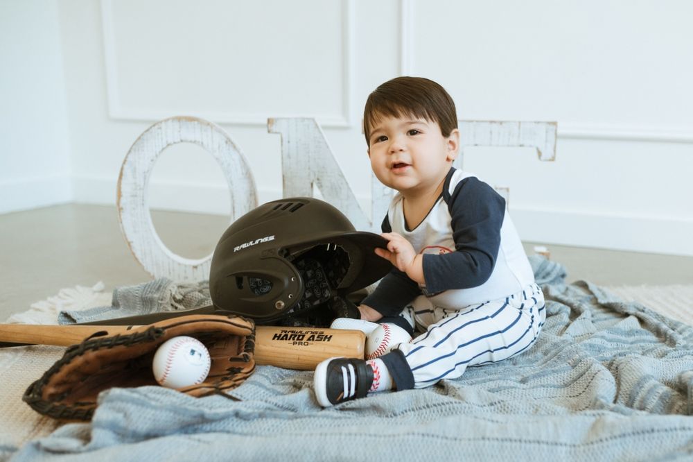 A young child sits on a bed beside baseball equipment including a helmet, bat, and glove in a bright room.