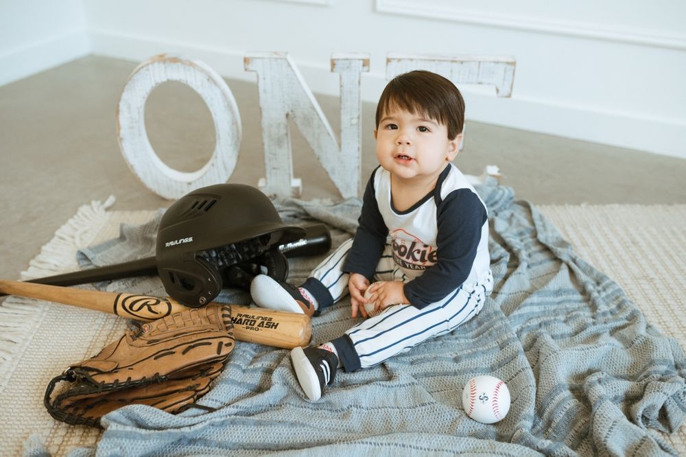 A young baseball fan sits on a bed with baseball gear including a bat, glove, and ball.