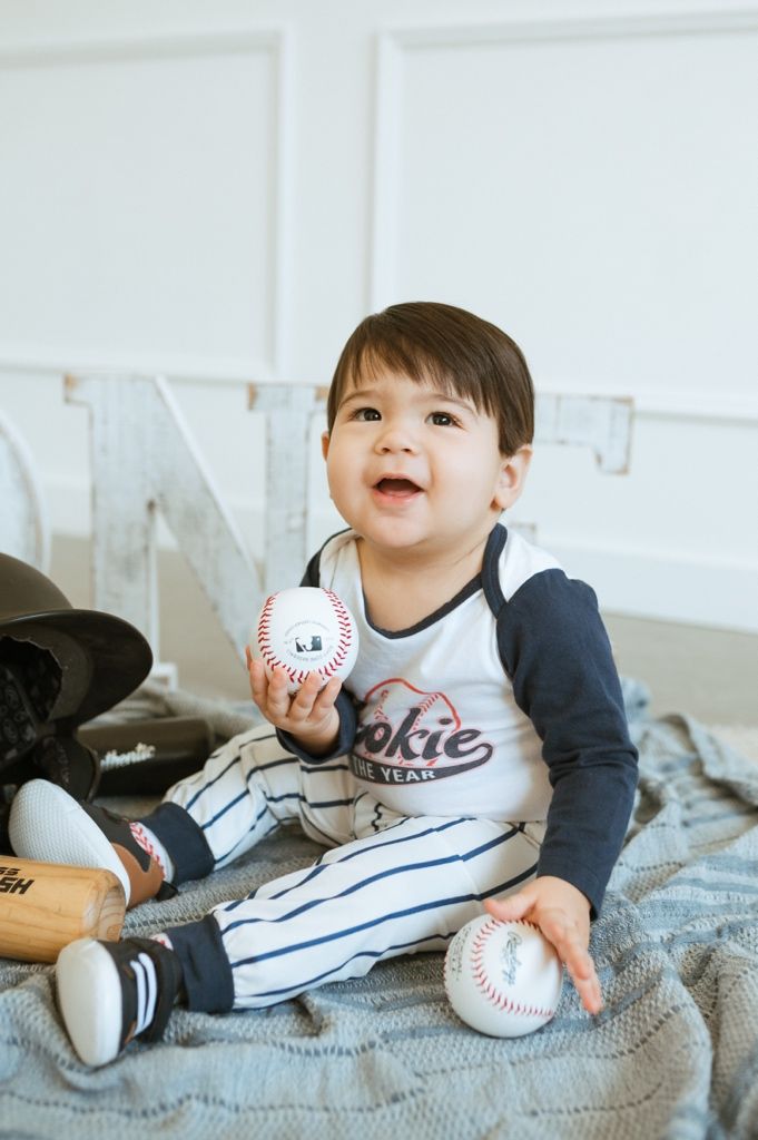 Toddler in baseball-themed outfit sits on gray blanket holding baseballs and smiling happily.