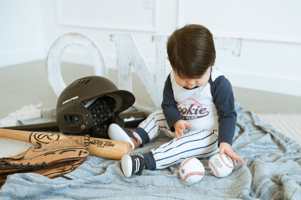 Small child sitting on bed reading with baseball equipment and gloves nearby.