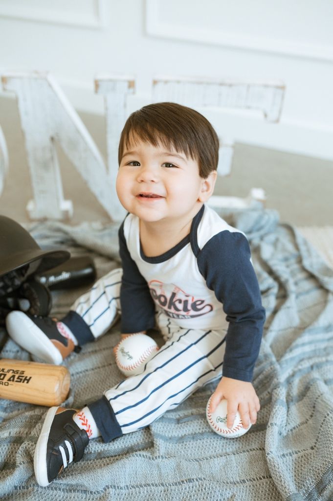 Small child wearing a navy sweater and white sneakers lying on a gray knit blanket with a playful happy expression.