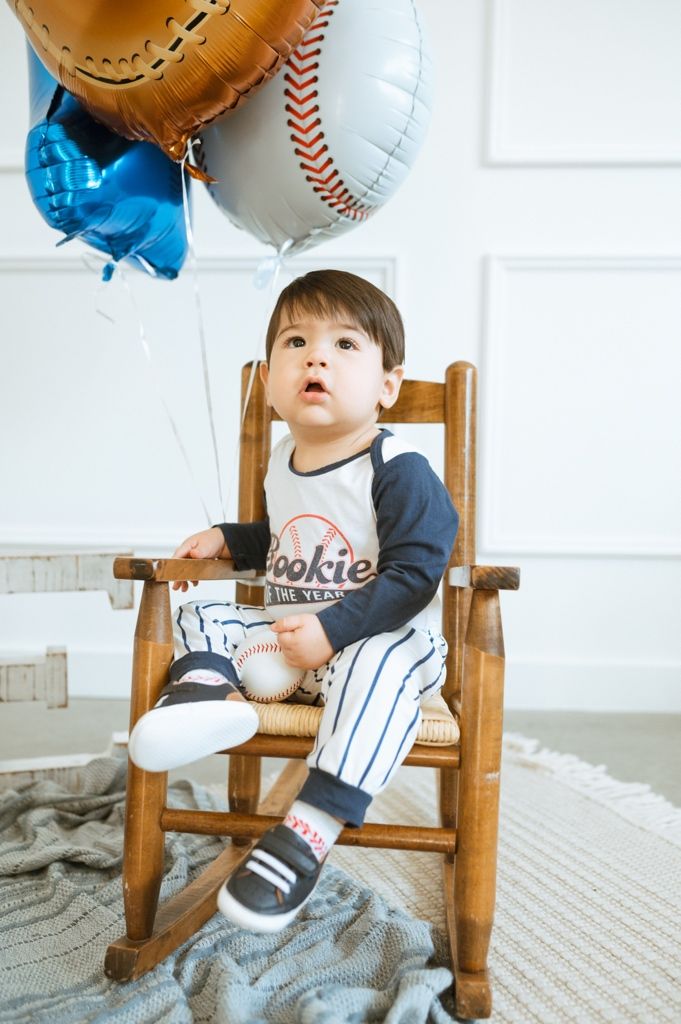 Small child sits in wooden rocking chair with baseball-themed balloons and decor for a special celebration.