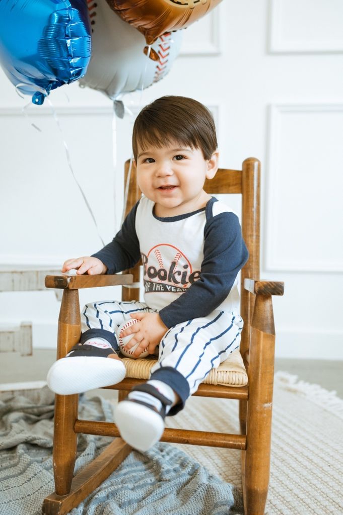 Young child sits in wooden rocking chair with baseball-themed decorations and balloons.