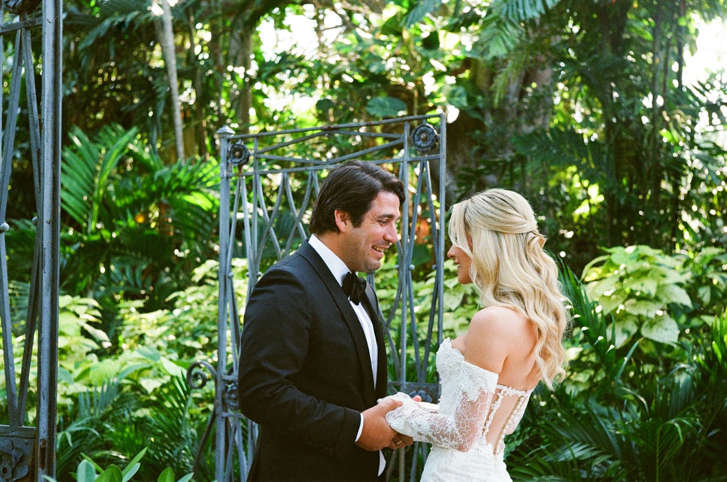Couple embraces in a lush garden setting during outdoor wedding ceremony at sunny venue decorated with white florals.