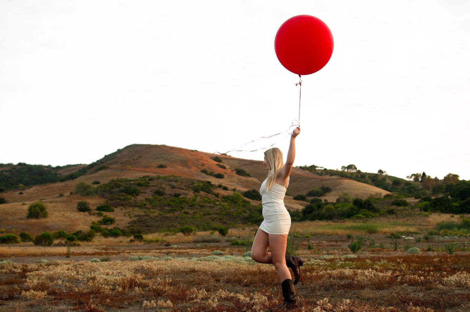 Blonde woman in white outfit holding a large red balloon in an open field with rolling hills.