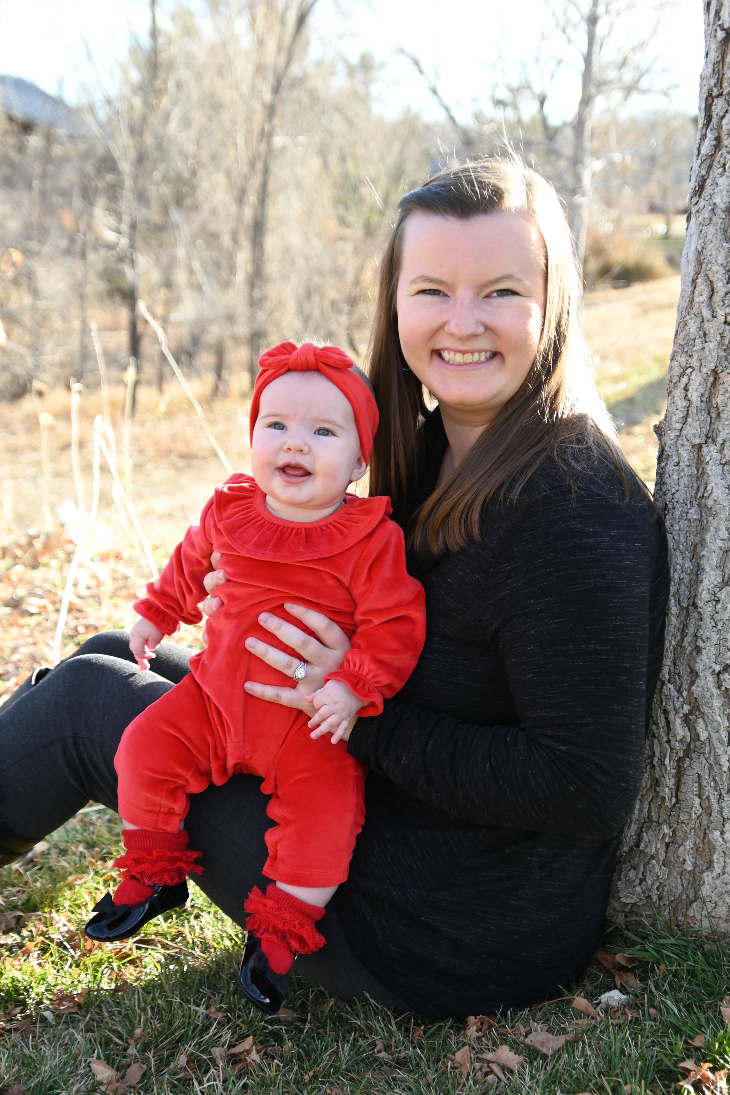 A family outdoor photoshoot shows sweet moments between parent and child in red sitting by a tree on autumn day.