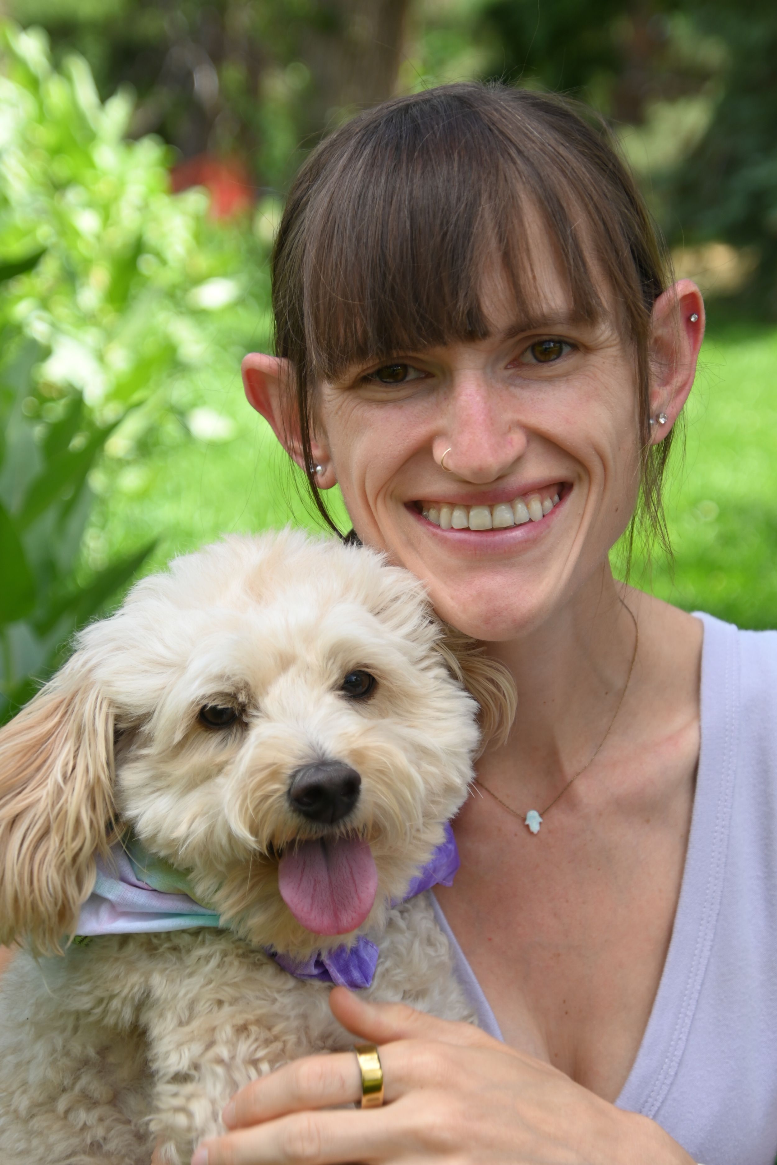 A cute fluffy white dog with a purple collar smiling with its owner in a sunny outdoor setting with green foliage.