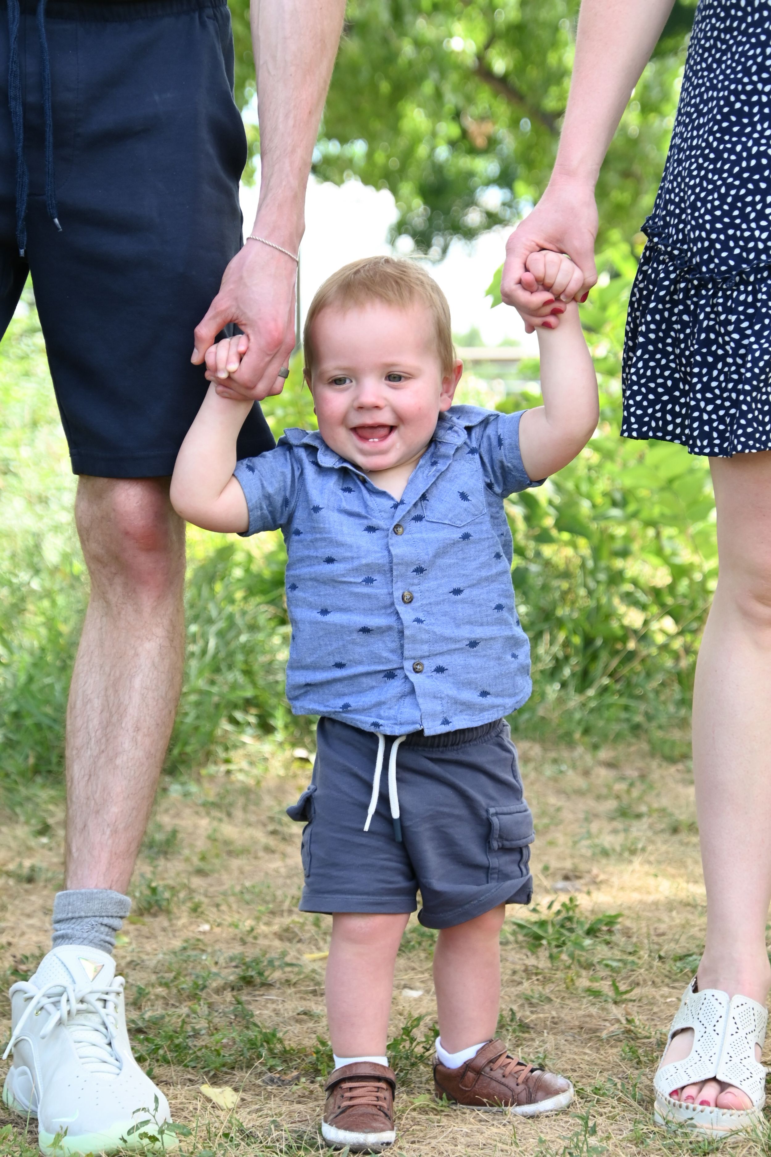 A cheerful toddler in a blue button-down shirt and shorts holds hands with adults while standing outdoors in summer.