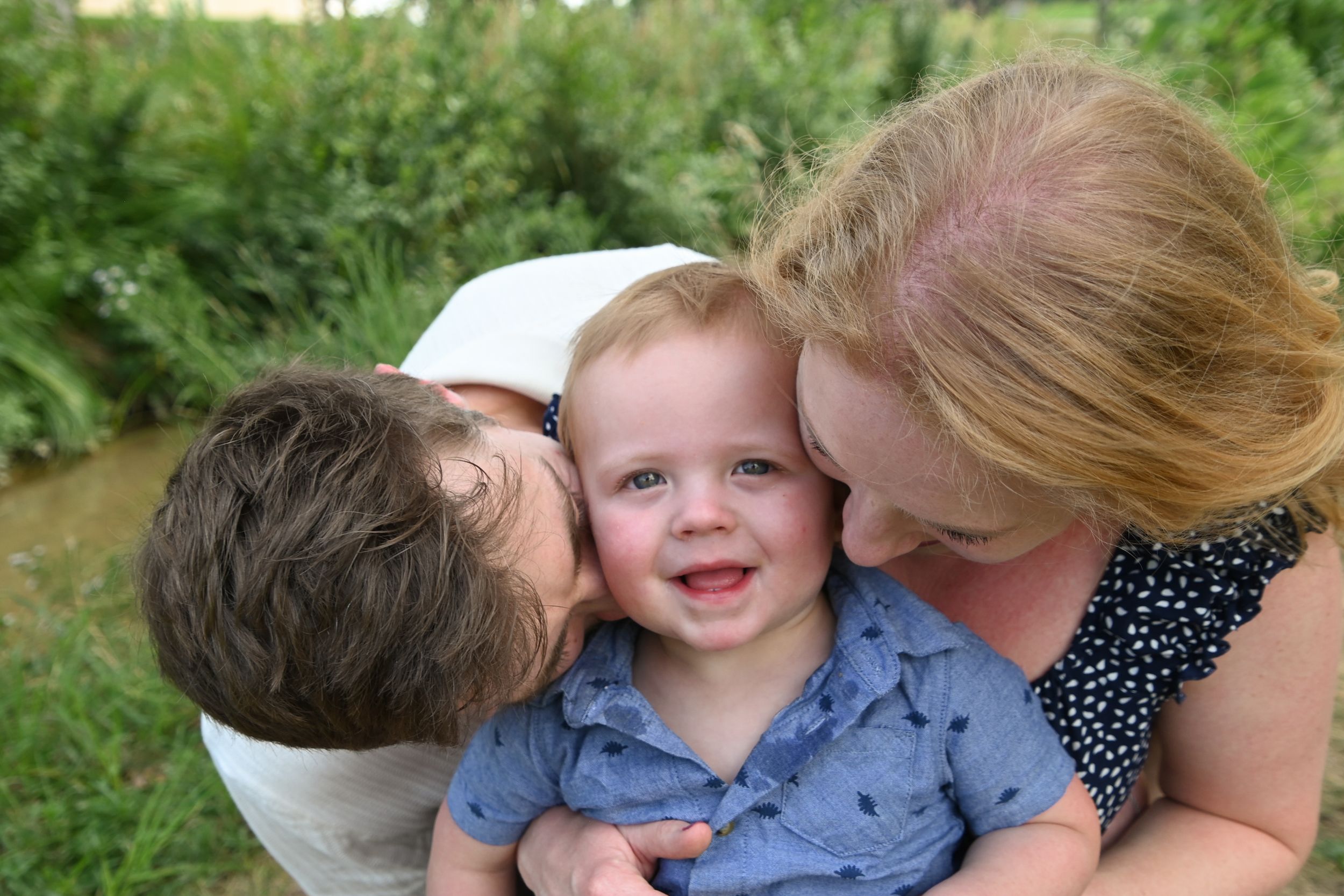 A tender family moment captured outdoors with loving embraces and smiles against a green natural background.
