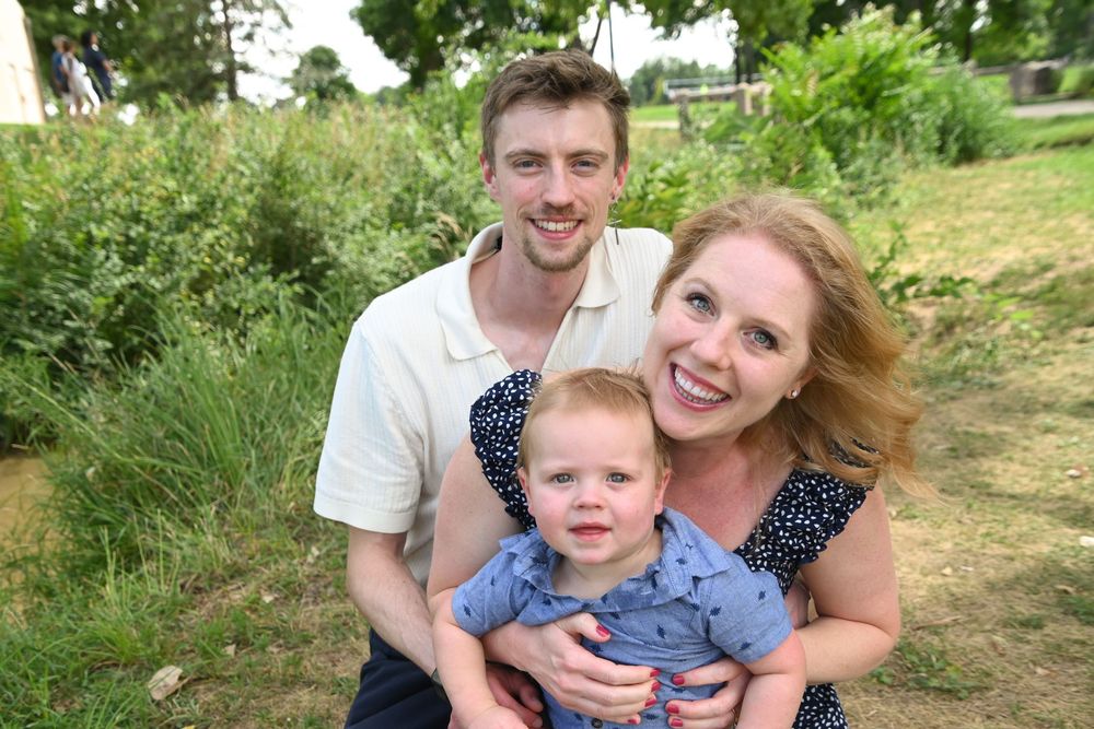 A family of three poses outdoors on a grassy hillside on a sunny summer day.
