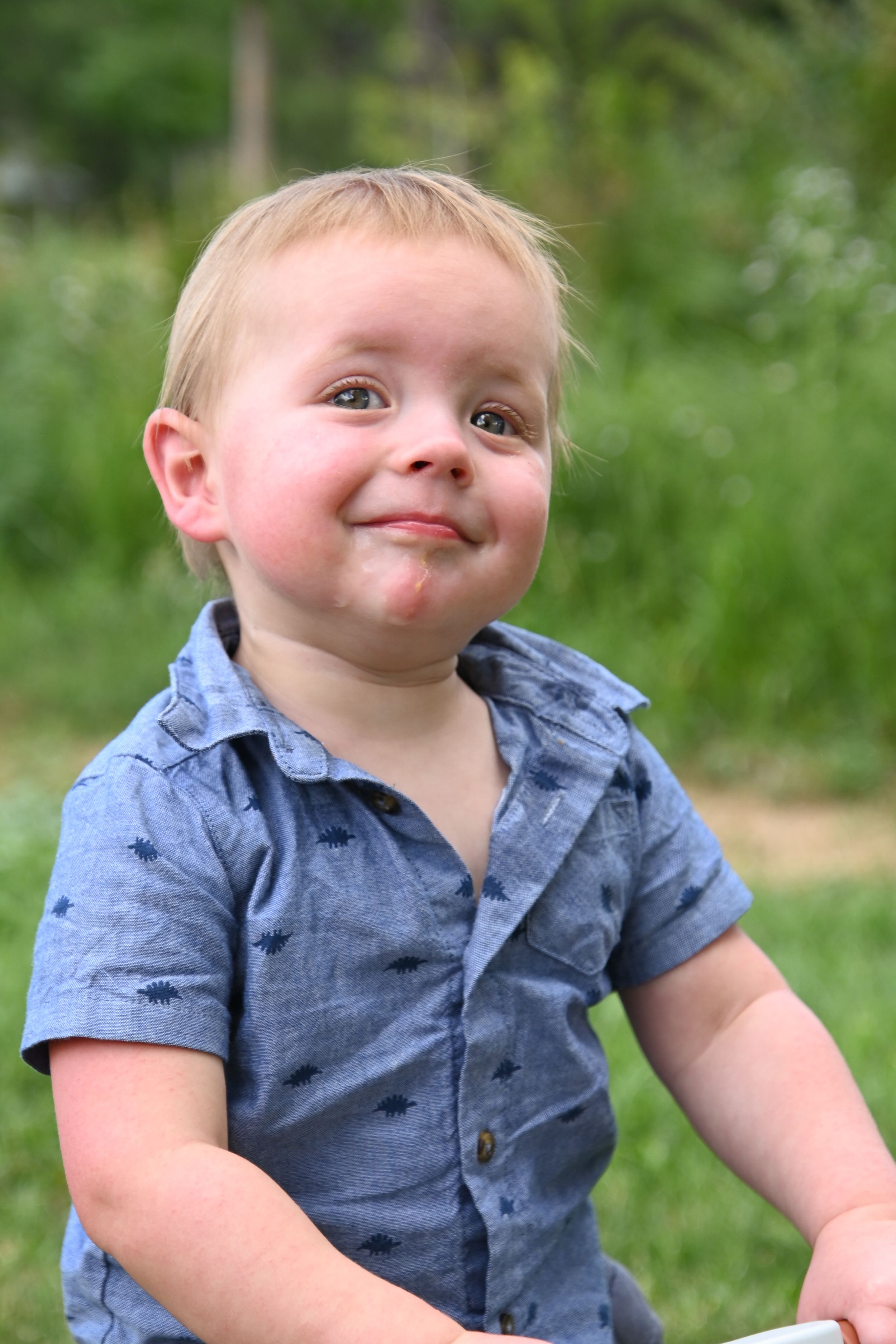 A small child wearing a blue chambray button-down shirt smiles playfully outdoors against a green background.