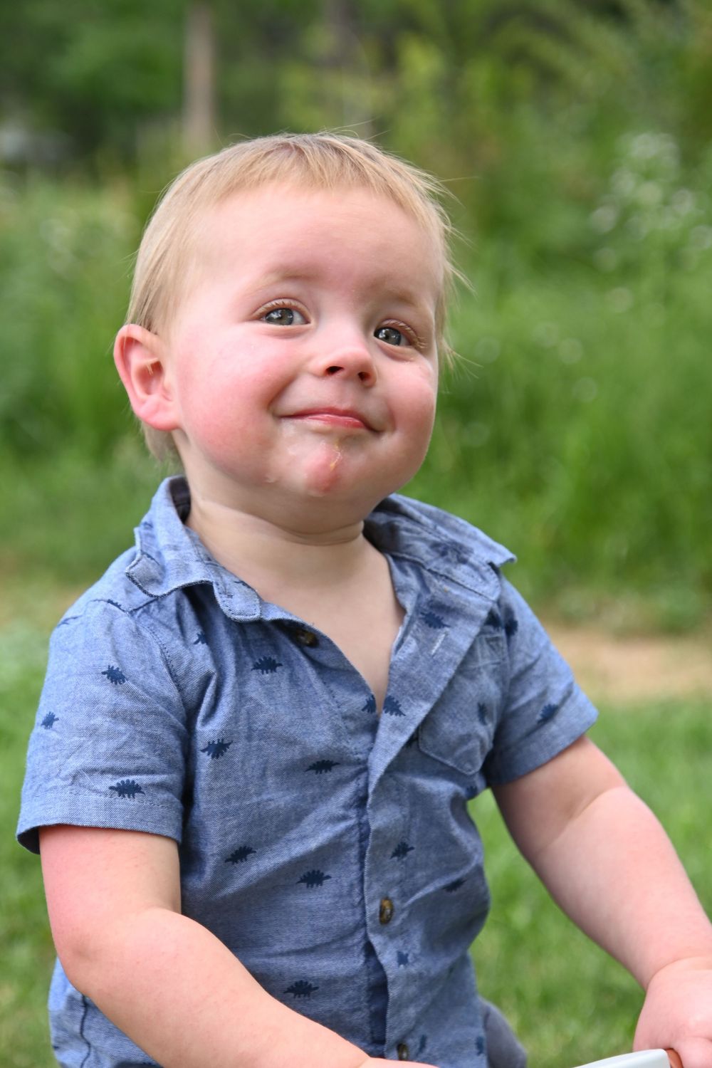 A small child wearing a blue chambray button-down shirt smiles playfully outdoors against a green background.
