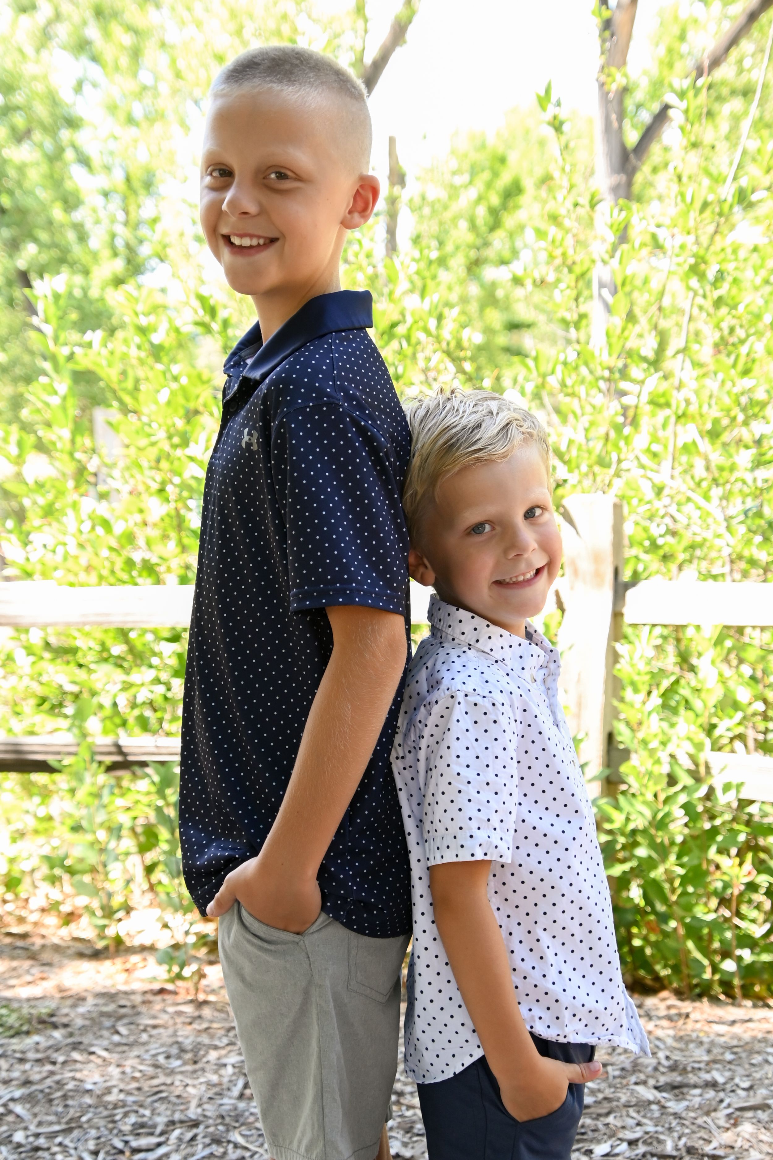 Two children wearing polka dot shirts stand back to back outdoors with bright green foliage in the background.