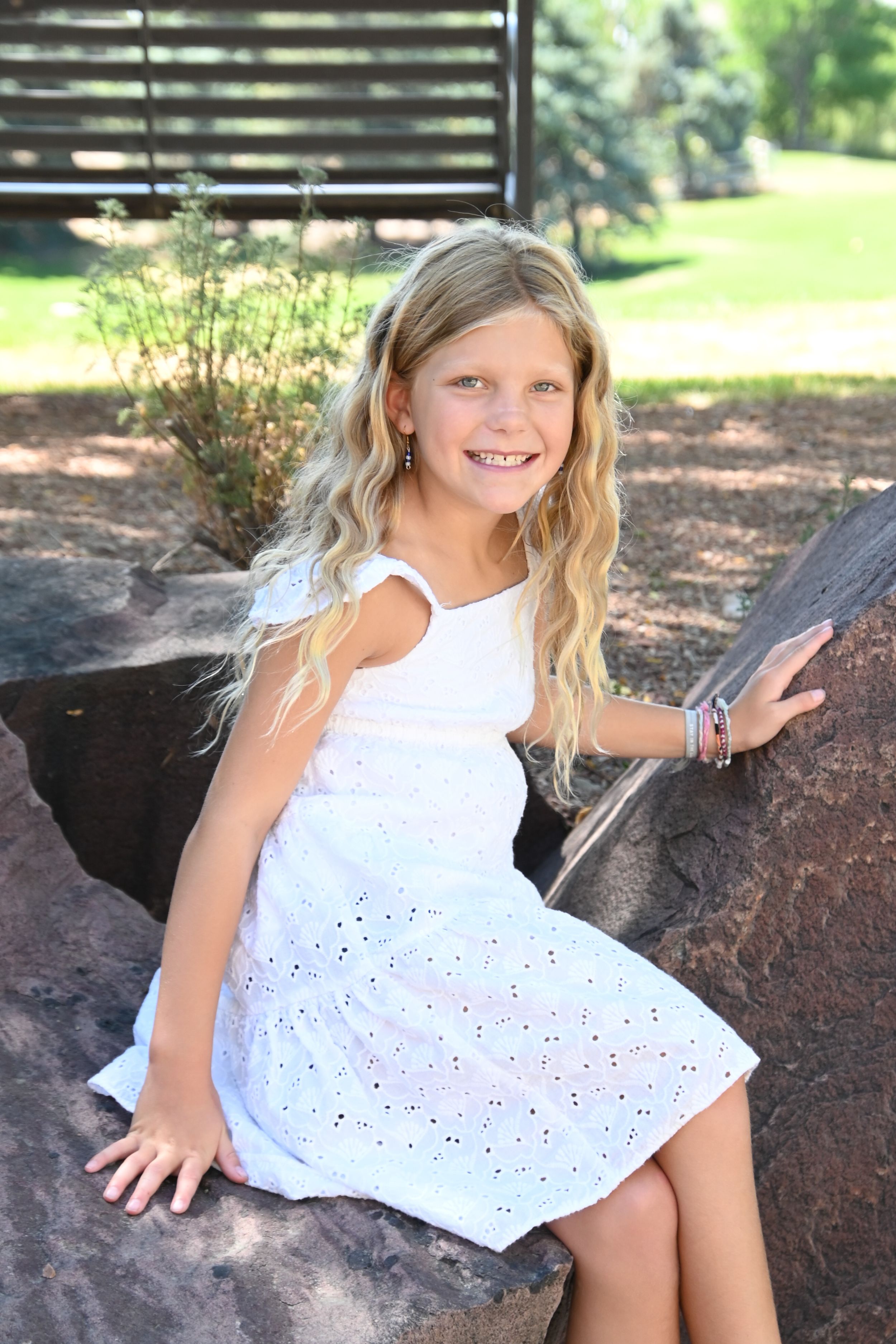 A young child in a white eyelet dress sits on a large rock in a sunny outdoor setting.
