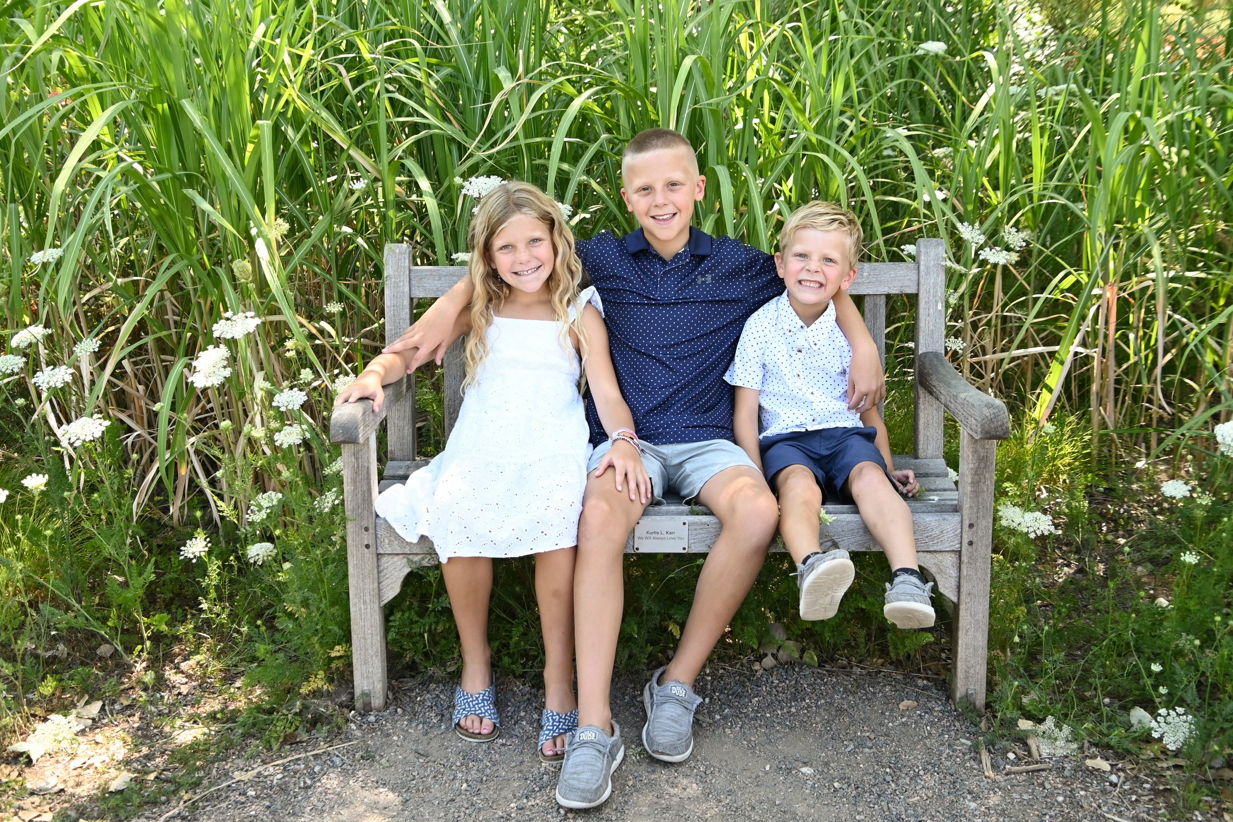 Smiling children sitting together on a wooden bench surrounded by tall green grass and wildflowers in summer.