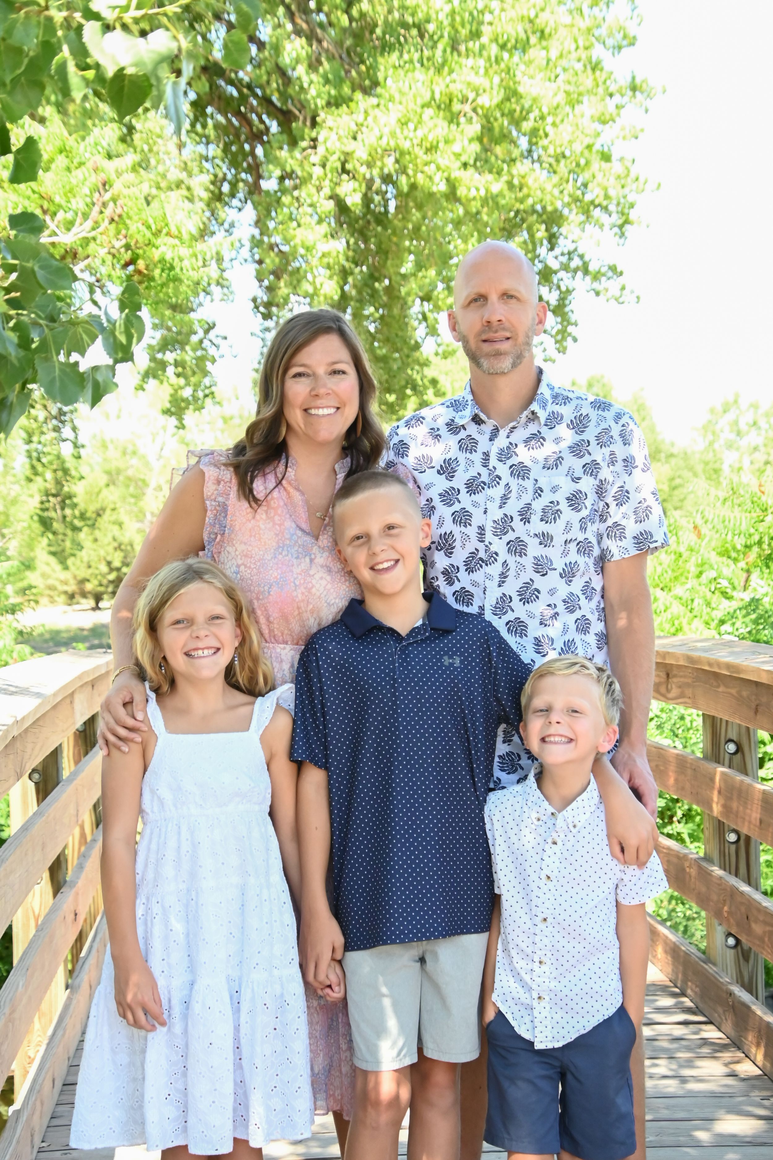 A family poses together on a wooden bridge in coordinated blue and white summer outfits in natural sunlight.