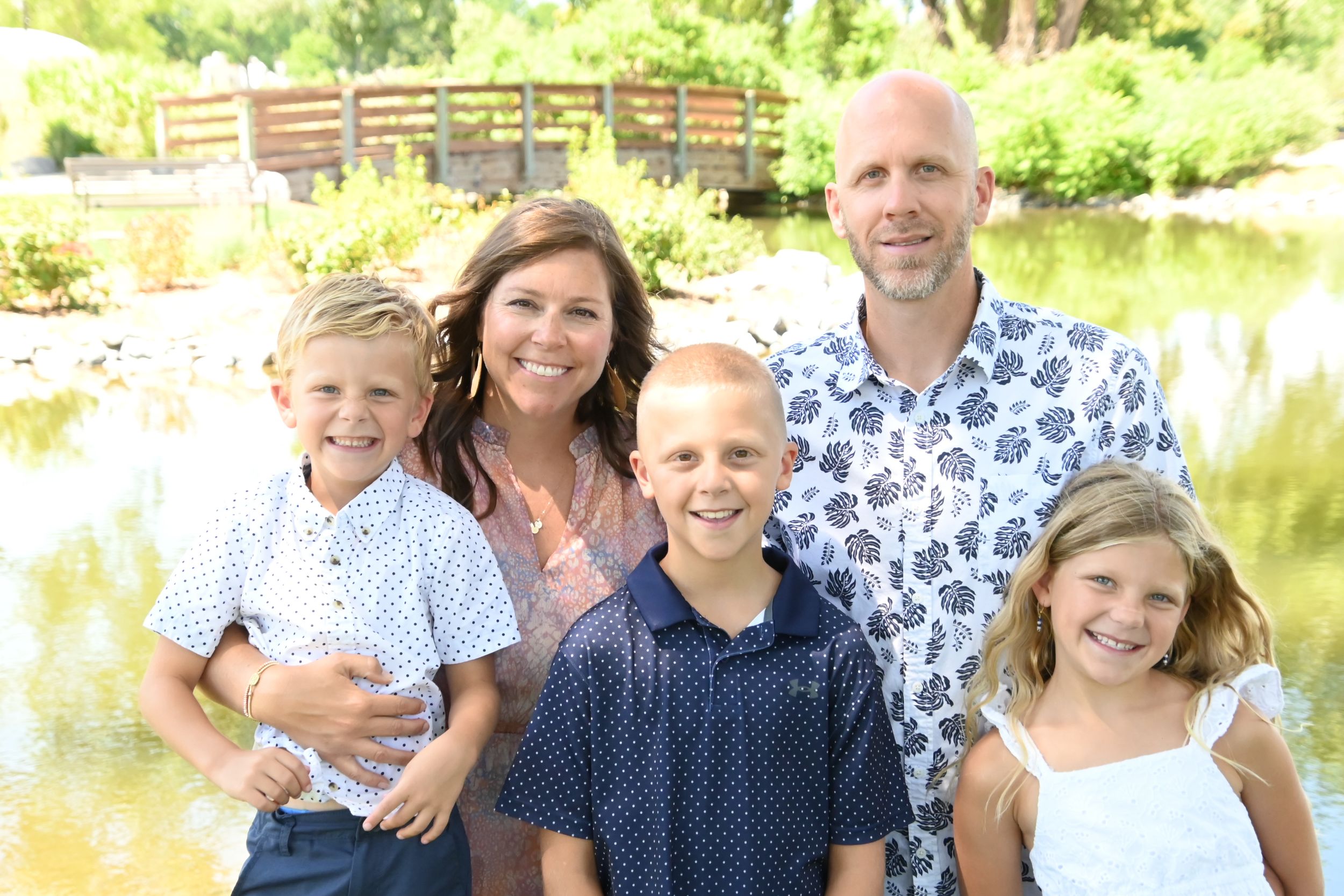 A family wearing coordinated blue and white outfits poses together outdoors near a lake on a sunny day.