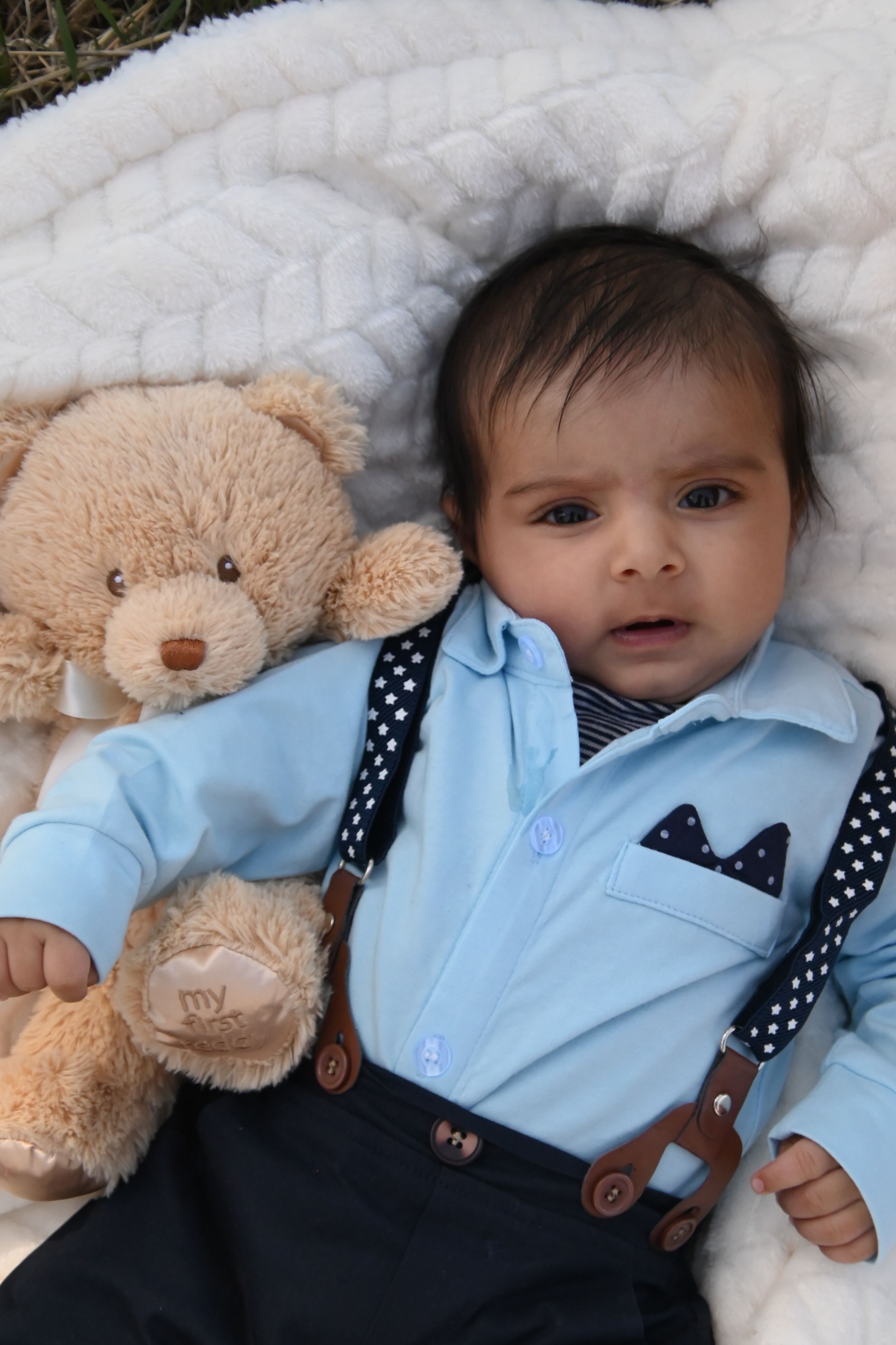Baby wearing light blue dress shirt and polka dot suspenders lies on white bedding next to plush teddy bear toy.